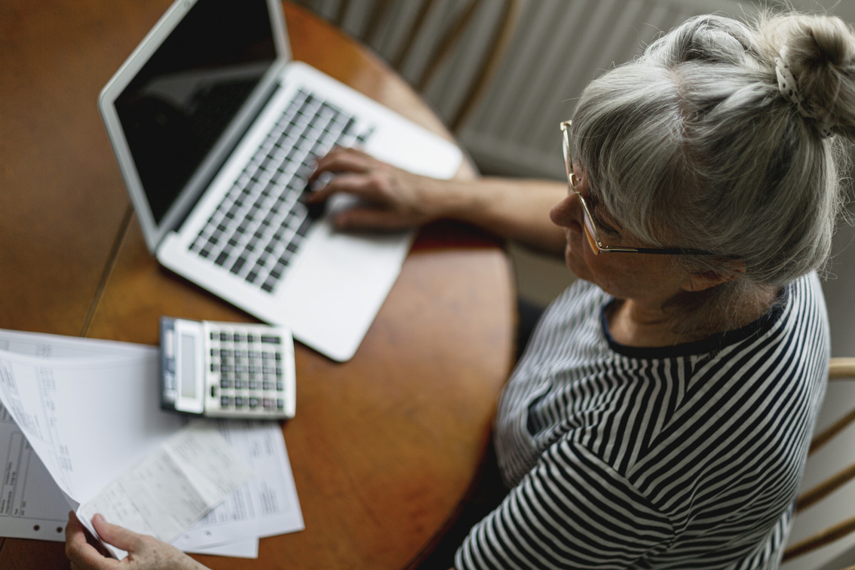 Eine ältere Frau mit grauen Haaren sitzt vor ihrem Laptop und schaut auf Rechnungen.