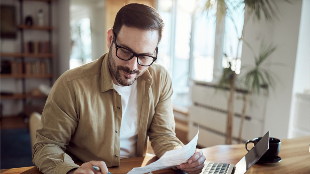 Ein Mann sitzt mit Stift, Papier und einem Rechner sm Schreibtisch und lächelt zufrieden.