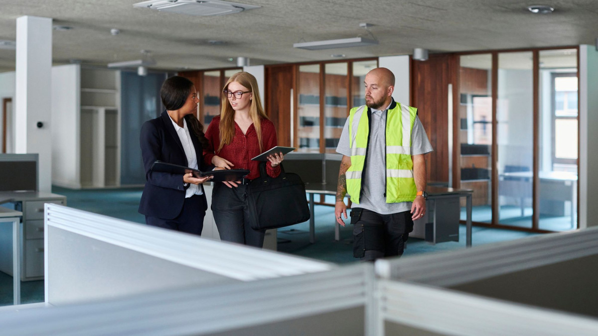 Zwei Frauen in Businesskleidung und ein Mann mit Warnweste gehen durch ein leeres Bürogebäude