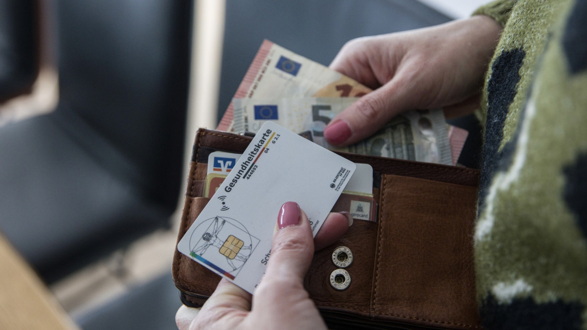 Eine Frau entnimmt Euro-Banknoten aus einem Portemonnaie, in dem sich neben Bargeld auch mehrere Zahlungskarten sowie eine elektronische Gesundheitskarte befinden