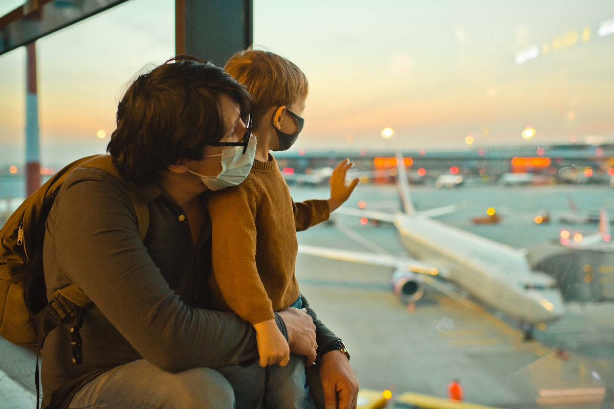 Ein Vater und ein Sohn mit Atemschutzmaske am Fenster eines Flughafens