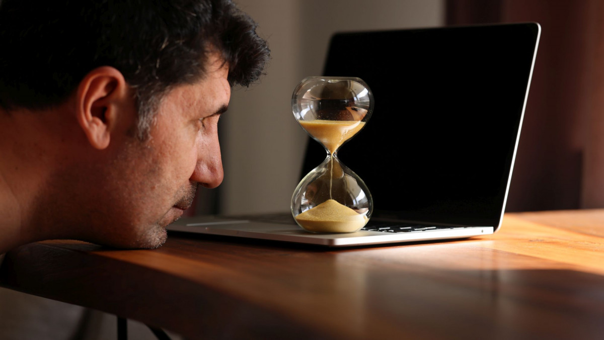 Mann schaut auf Sanduhr vor Laptop