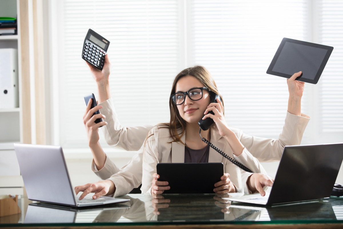 Eine Frau hat sechs Hände und hält in jedem ein anderes Device wie einen Laptop, ein Telefon, ein Handy etc