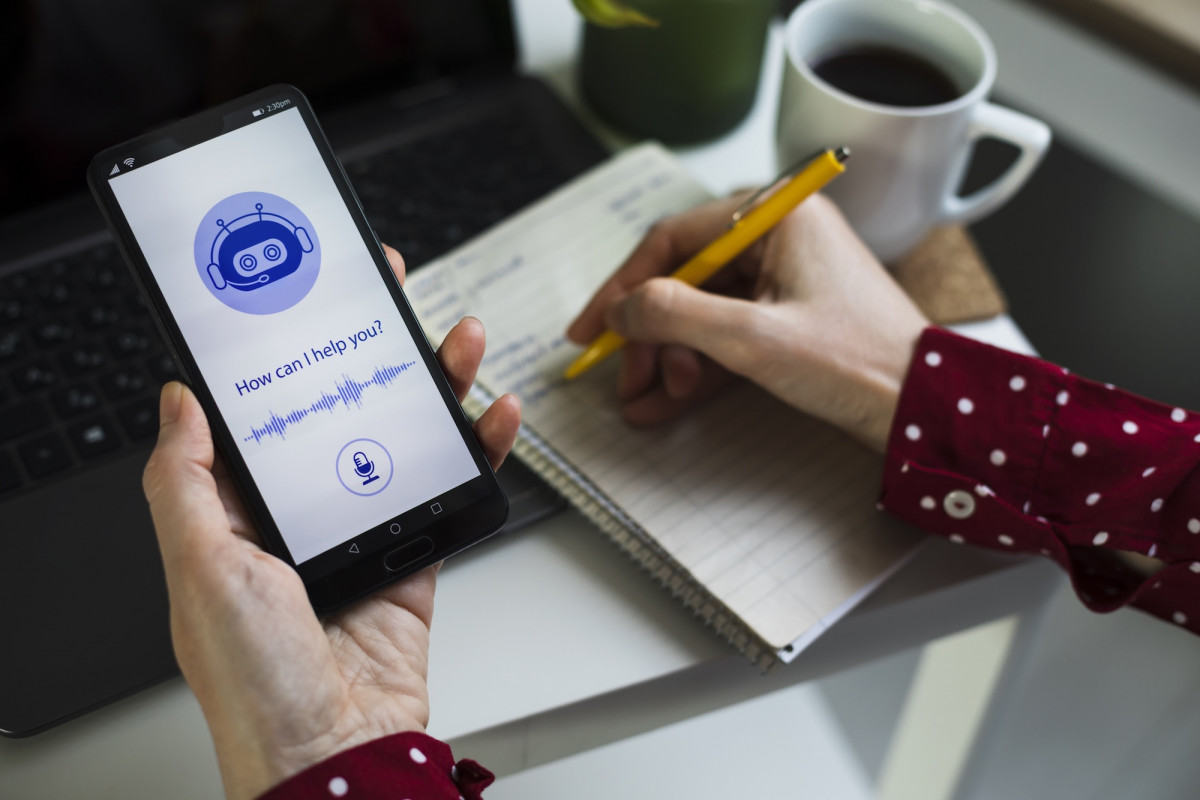 Ein Handy in der einen Hand einer Frau, in der anderen Hand hält die Frau einen Stift und schreibt in einen Block. Auf dem Display des Handys steht: How can i help you?