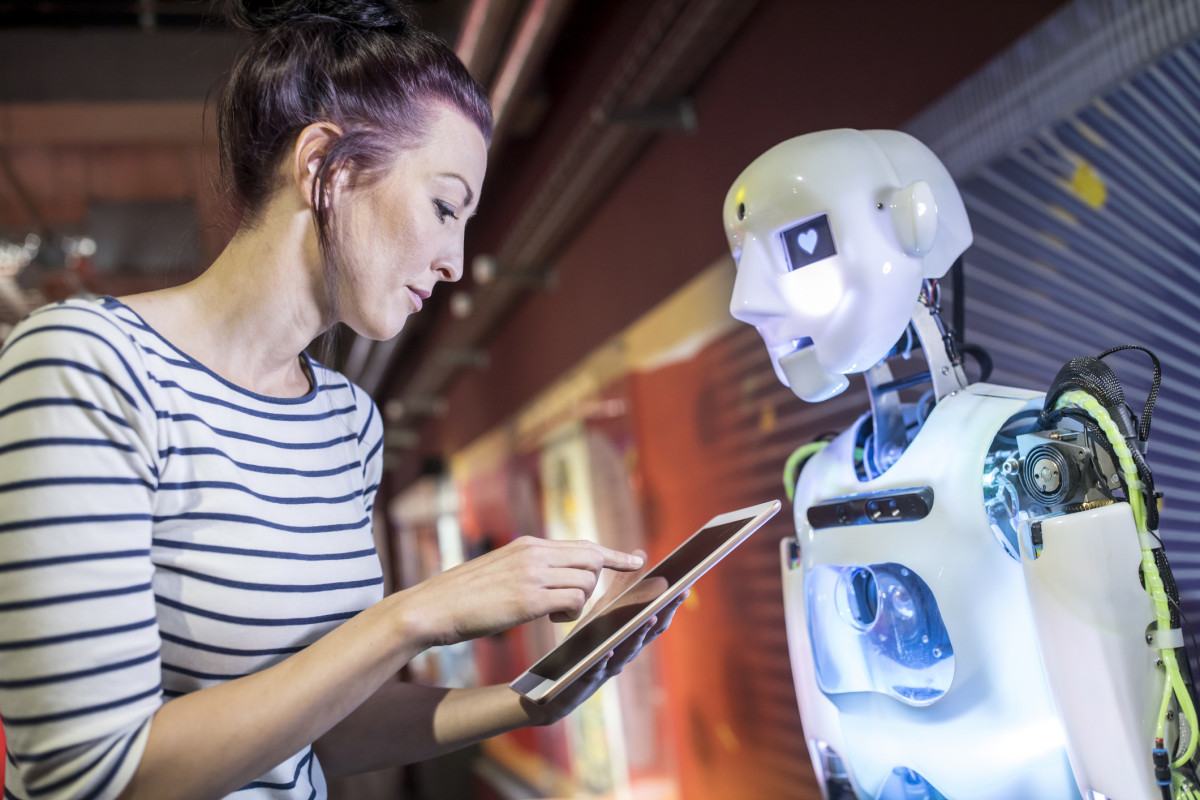 Eine Frau und ein Roboter stehen sich gegenüber. Die Frau hält ein Tablet und im Auge des Roboters ist ein Herz zu sehen.