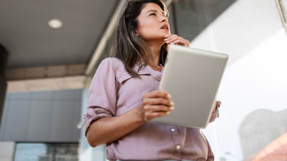 Eine Frau hält ein iPad in der Hand und schaut nachdenklich in die Ferne.