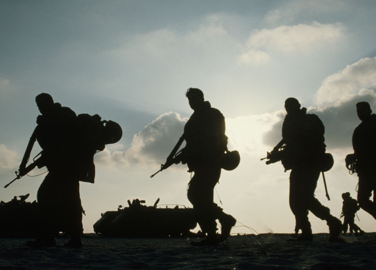 Ein Gruppe Soldaten marschiert. Im Hintergrund sieht man gepanzerte Fahrzeuge.