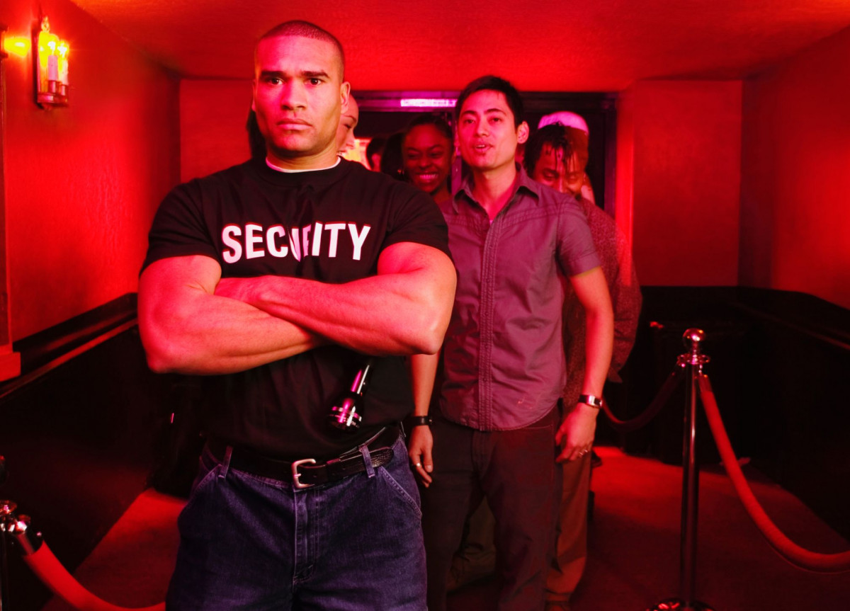 Ein Türsteher steht grimmig vor einem Club, hinter ihm sind einige feiernde Menschen