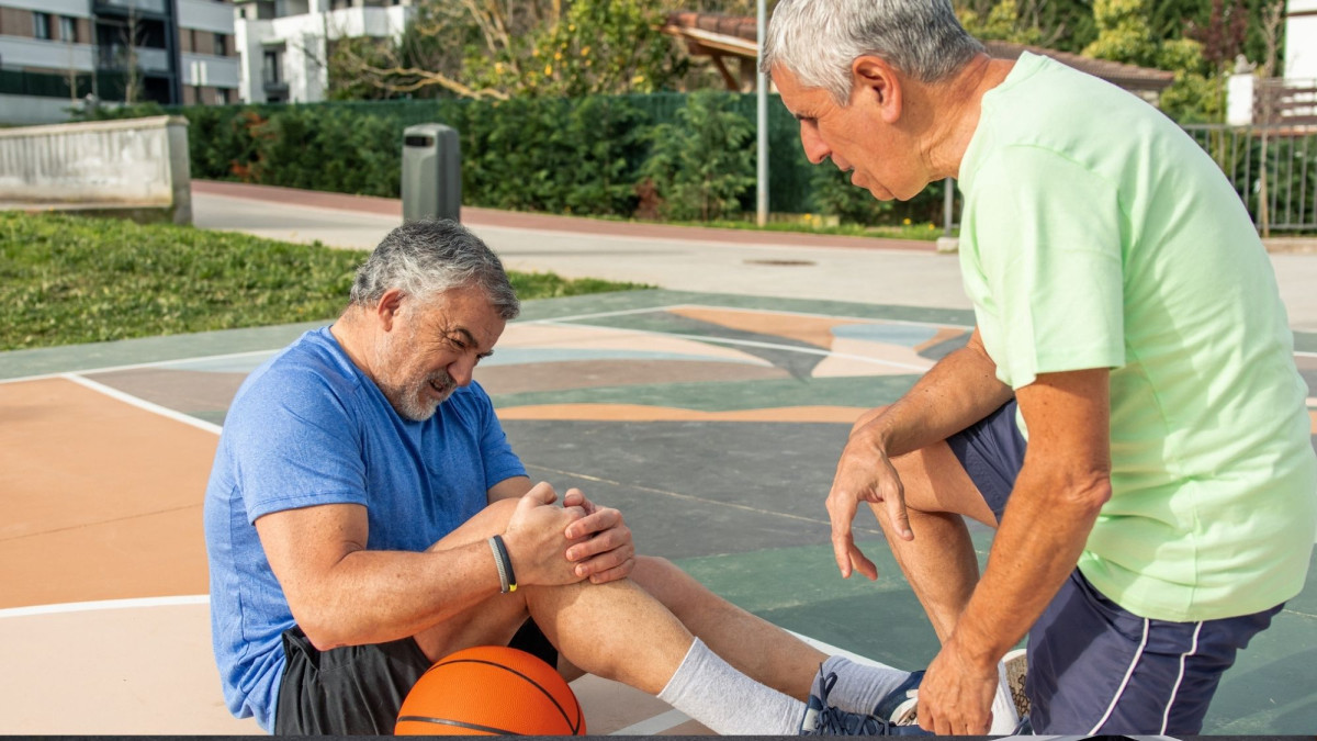Zwei ältere Männer beim Basketball. Der eine sitzt auf dem Boden und hält sich das Knie, der andere kniet vor ihm und guckt sorgenvoll.