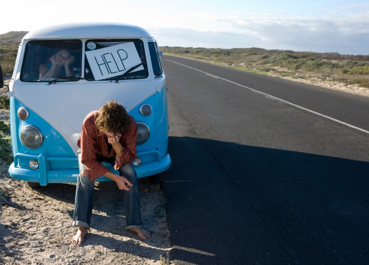 Ein Mann sitzt mit hängendem Kopf vor einem liegengebliebenen VW-Bus, in der Scheibe steckt ein Zeichen mit der Aufschrift "Help"